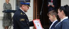 Komendant miejski PSP wręcza chłopcu odznakę i dyplom komendanta główngo PSP. Obok stoi mama chłopca, w tle budynek szkoły i dwie osoby z obsługi.