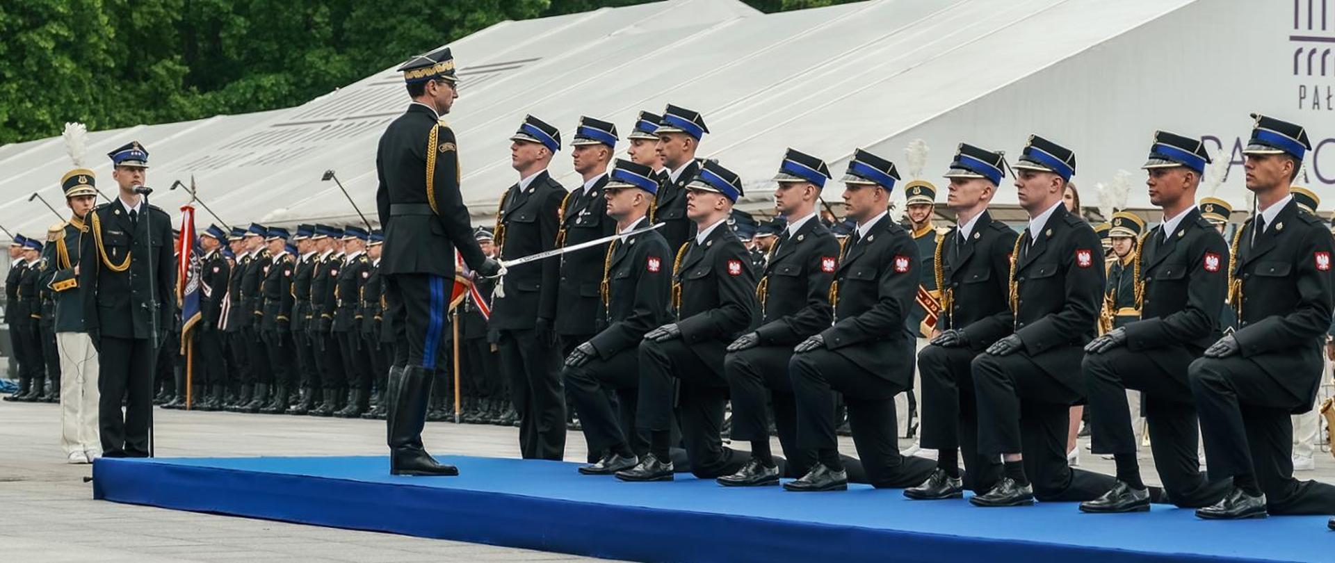Oficjalne uroczystości państwowe. Na pierwszym planie, na długim podwyższeniu pokrytym niebieskim materiałem, klęczy rząd strażaków w czarnych mundurach galowych, czapkach rogatywkach i rękawiczkach. Przed nimi stoi oficer w mundurze - generał, który dokonuje aktu promowania szablą. W tle widać rzędy innych pododdziałów mundurowych, białe namioty halowe oraz fragment napisu na budynku.