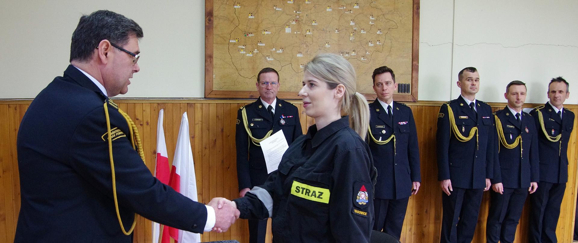Widok z przodu. Po lewej stronie komendant powiatowy PSP ściska dłoń stojącej naprzeciwko str. Annie Janeczko. Na drugim planie kadra KP PSP w Wadowicach.