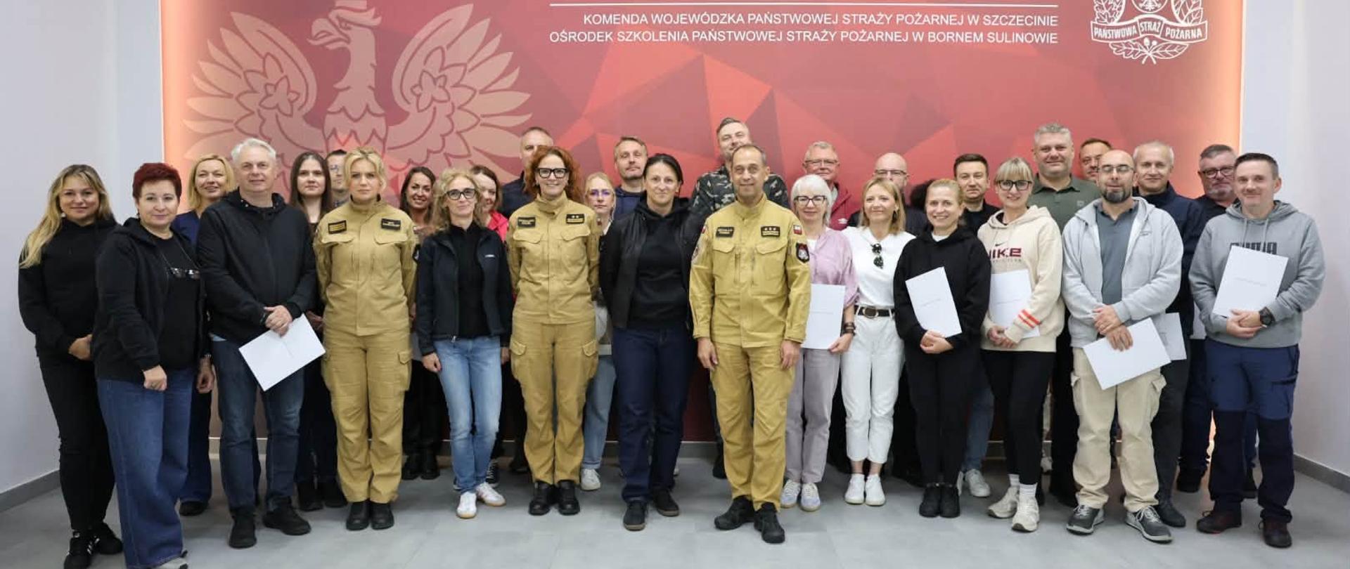 W sali szkoleniowej Ośrodka Szkolenia Państwowej Straży Pożarnej w Bornem Sulinowie stoi grupa uczestników szkolenia. W centrum znajdują się strażacy w umundurowaniu, a wokół nich pozostali uczestnicy w ubraniach codziennych. Część osób trzyma białe dokumenty. W tle znajduje się duża ściana z logo Państwowej Straży Pożarnej oraz napisem „Państwowa Straż Pożarna – Komenda Wojewódzka Państwowej Straży Pożarnej w Szczecinie – Ośrodek Szkolenia Państwowej Straży Pożarnej w Bornem Sulinowie”.