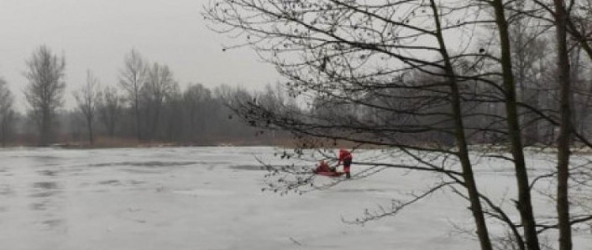 Zamarznięte jezioro. Na środku widoczna para strażaków w ubraniach do pracy w wodzie. Wyposażeni w sanie lodowe przemieszczają się po tafli lodu w kierunku osoby poszkodowanej.