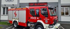 Na zdjęciu widzimy samochód pożarniczy koloru czerwonego, marki Renault. Samochód stoi bokiem do kadru, widzimy żaluzje od skrytek koloru srebrnego. Na przednich drzwiach samochodu widzimy herb gminy jednak nie można odczytać jakiej oraz biały napis OSP Blizanów. Na tylnych drzwiach samochodu biały numer operacyjny: trzysta czterdzieści dziewięć p dwadzieścia osiem. Na dachu samochodu widoczne są sygnały świetlne oraz węże ssawne koloru zielonego. Samochód usytuowany jest przed budynkiem Ochotniczej Straży Pożarnej w Blizanowie, stoi na asfalcie koloru szarego. W tle widzimy budynek remizy OSP Blizanów, dwukondygnacyjny koloru białego na ścianie zawieszony szyld z napisem OSP BLIZANÓW koloru czerwonego.