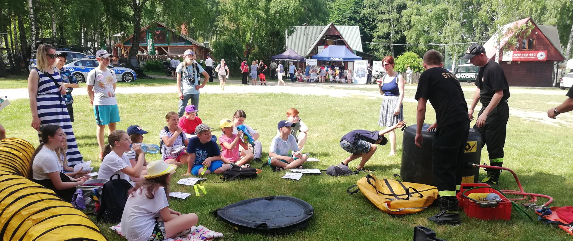 Na zdjęciu widzimy siedzące dzieci którym dwóch funkcjonariuszy prezentuje działanie sprzętu używanego do działań ratowniczo-gaśniczych w jednostce KP PSP Lesko oraz dzieci Na Drugim planie widzimy dzieci znajdujące się pod namiotem policji oraz samochody policyjne. W tle znajdują się budynki Bieszczadzkiego Wodnego Ochotniczego Pogotowia Ratunkowego oraz budynek restauracji.