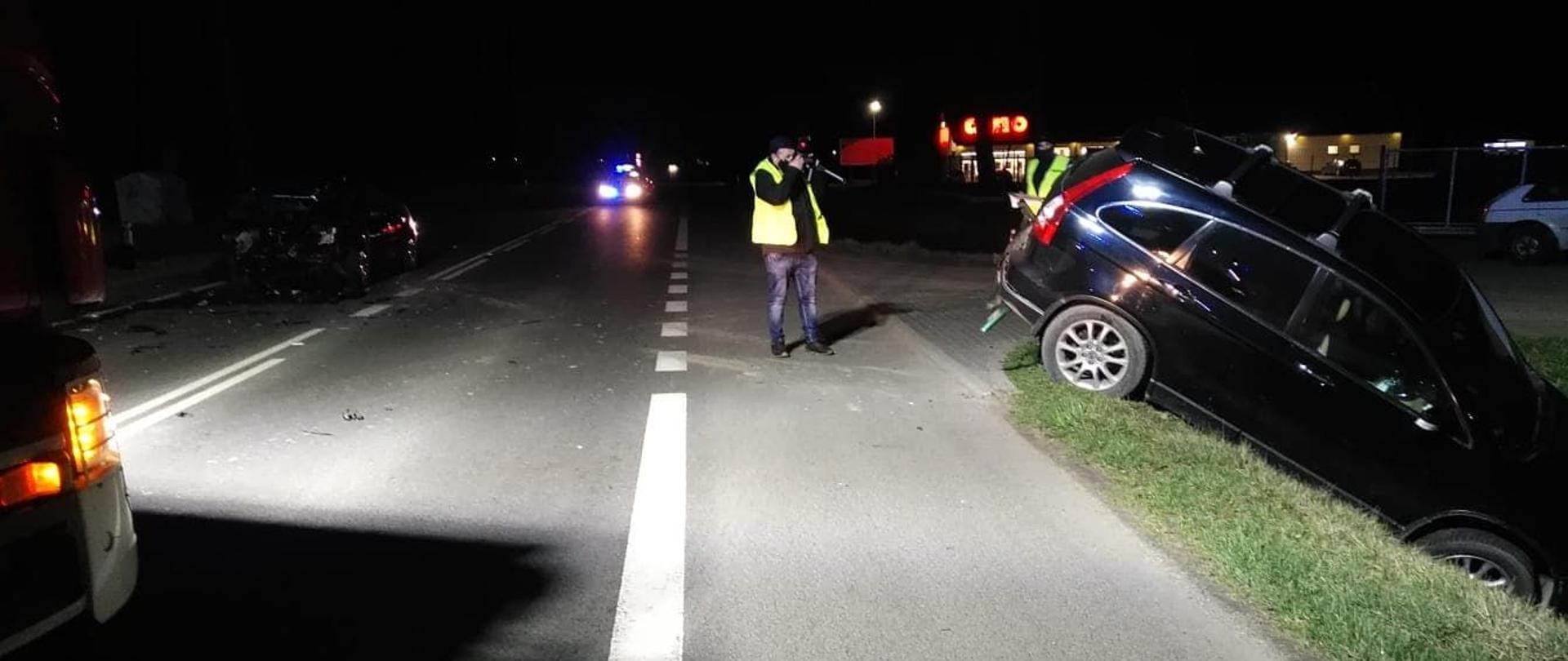Zdjęcie przedstawia rozbity samochód który z powodu uderzenia przez inny samochód wjechał do rowu przy drodze. Na drodze stoi policjant który robi zdjęcia zdarzenia.jest pora nocna.