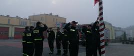 Strażacy stoją w dwóch rzędach naprzeciwko siebie oddając honor poczet sztandarowy wciąga flagę na maszt za nimi stoją pojazdy pożarnicze. 