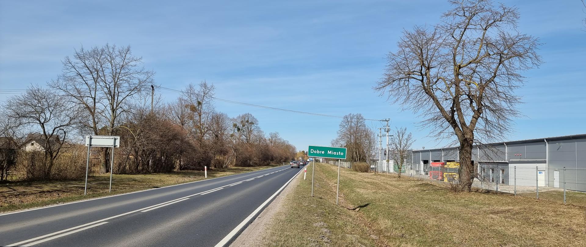 Asfaltowa droga na przedmieściach. W oddali sznur samochodów i tabliczka z nazwą miasta.