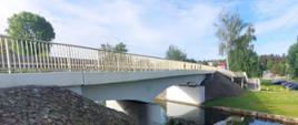Most nad zbiornikiem wodnym, z poręczami po obu stronach. Most jest podtrzymywany przez betonowe filary i prowadzi do niego kamienny nasyp. Wokół znajduje się zieleń, w tym drzewa i trawa. Na niebie widać kilka chmur.