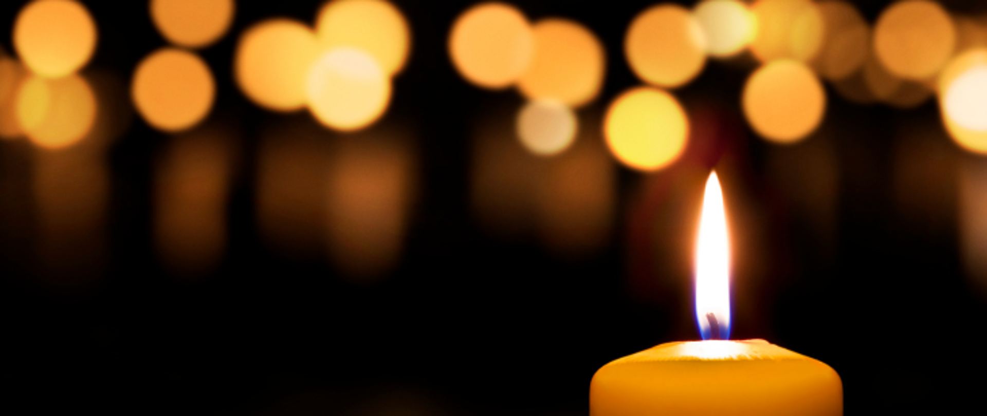 Na ciemnym tle u dołu z prawej strony znajduje się zapalona świeca. U góry znajdują się odblaski innych świec.