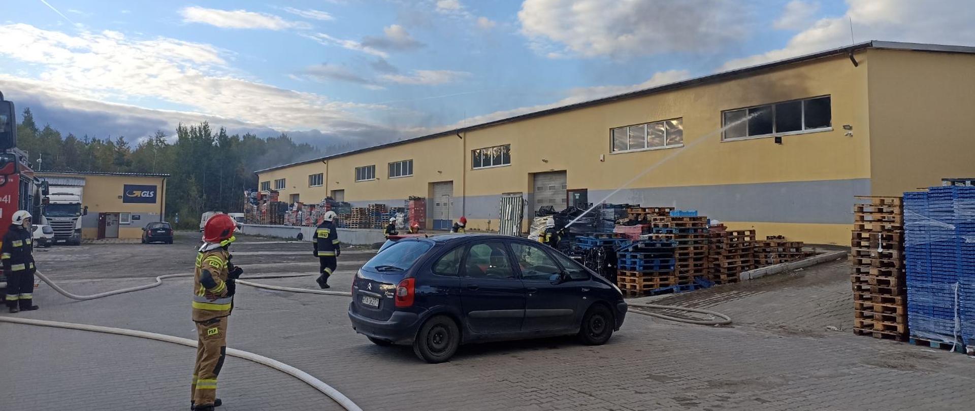 Na placu przed budynkiem magazynowym trwa akcja gaśnicza, strażacy gaszą pożar wewnątrz budynku podając wodę przez okna. Z okien wydobywa się dym. Przed budynkiem ustawione są różnego rodzaju palety oraz opakowania. 