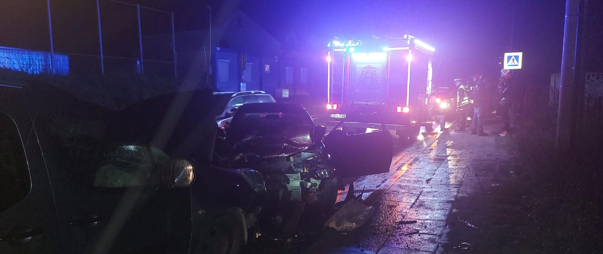 Zdjęcie przedstawia wypadek trzech samochodów osobowych. Jest ciemno, zdjęcie jest rozświetlone niebieską poświatą pochodzącą od sygnałów alarmowych. Jezdnia i chodnik są mokre.