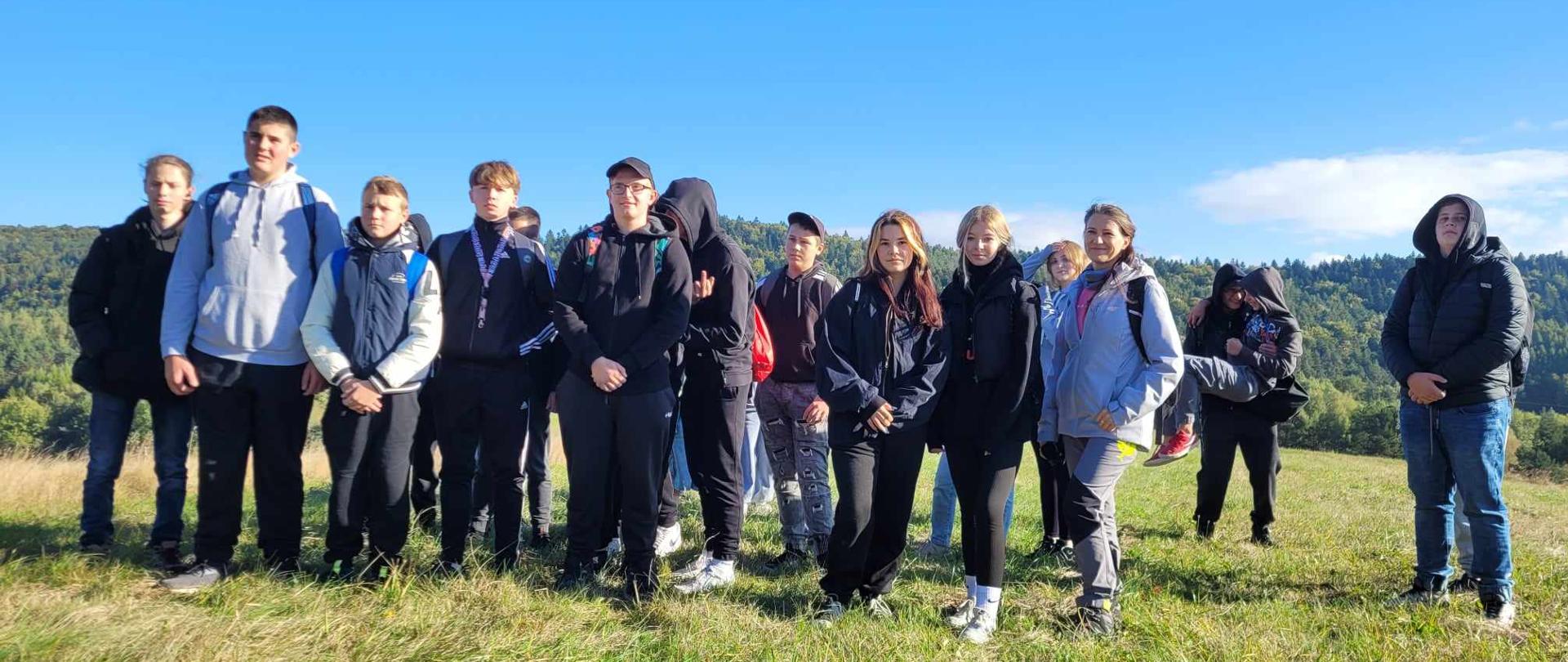 Portret grupowy młodzieży stojącej na szczycie wzniesienia. Wszyscy są ubrani w stroje sportowe i turystyczne, w tle rozciąga się widok na góry.