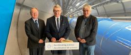 Minister Murdzek i dwóch mężczyzn w garniturach stoją za stojącą na słupku tabliczką z napisem Accelerating science, za nimi widać długi betonowy tunel w którym biegnie kilka rur.
