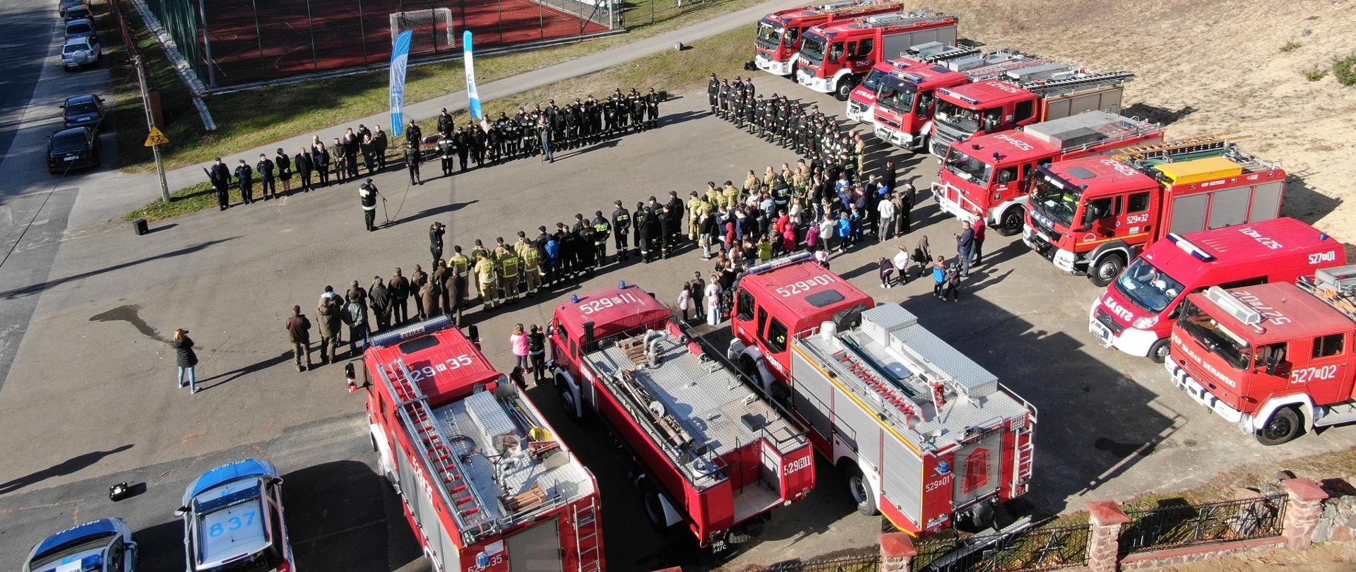 Fotografia przestawia miejsce zbiórki osób biorących udział w ćwiczeniach. Zdjęcie zostało wykonane z drona. Miejsce zbiórki znajduje się na placu odgrodzonym od boiska sportowego i budynków wąską drogą. Na pierwszym planie znajduje się wspomniany plac, na którym ustawione są samochody Państwowej Straży Pożarnej oraz samochody jednostek Ochotniczej Straży Pożarnej. Na placu widocznych jest łącznie 12 samochodów ustawionych na kształt odwrócoenj litery "L". W lewym dolnym rogu fotografii obok
samochodów PSP znajdują się dwa policyjne radiowozy. Przed samochodami ustawione są wszystkie osoby biorące udział w ćwiczeniach oraz inni obserwatorzy wysłuchujący
przemówienia. W dalszej części zdjęcia za drogą znajduje się ogrodzone siatką boisko sportowe oraz przyległe do niego budynki. Za budynkami i po prawej stronie zdjęcia
widoczne są drzewa.