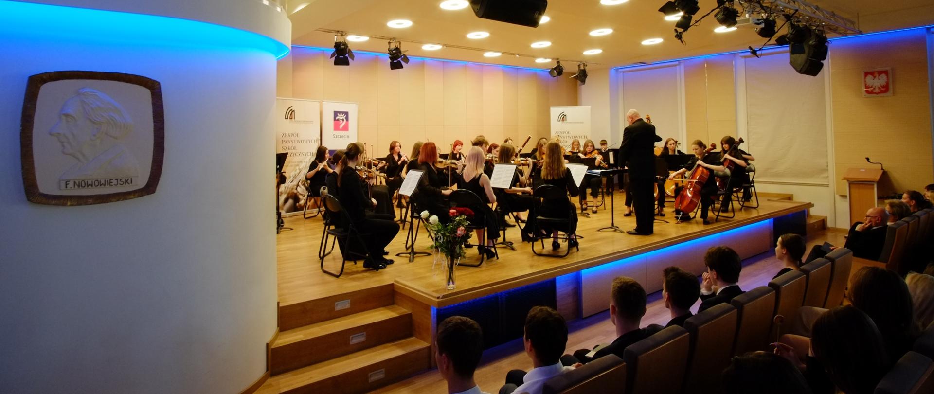 zdjęcie przedstawia orkiestrę szkoły, w ujęciu widoczne godło państwowe oraz płaskorzeźba z wizerunkiem patrona szkoły Feliksa Nowowiejskiego, w tle banery szkoły oraz miasta Szczecin