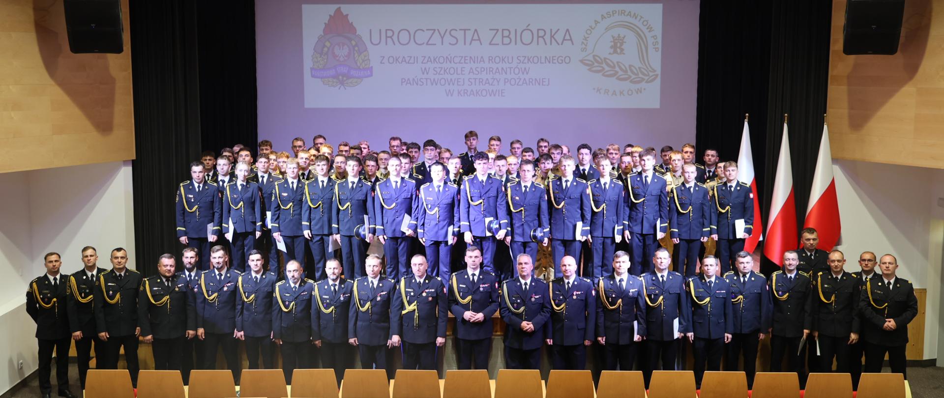  Oficjalne zdjęcie grupowe uczestników zbiórki
Szerokie ujęcie kadry i kadetów stojących w równych rzędach na scenie. UDuża grupa strażaków w ciemnogranatowych mundurach stoi pod ekranem z napisem „Uroczysta Zbiórka...”. Z przodu stoi kadra dowódcza ze sznurami galowymi, za nimi kadeci, a po prawej widać polskie flagi.

