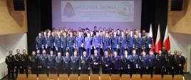 Oficjalne zdjęcie grupowe uczestników zbiórki
Szerokie ujęcie kadry i kadetów stojących w równych rzędach na scenie. UDuża grupa strażaków w ciemnogranatowych mundurach stoi pod ekranem z napisem „Uroczysta Zbiórka...”. Z przodu stoi kadra dowódcza ze sznurami galowymi, za nimi kadeci, a po prawej widać polskie flagi.
