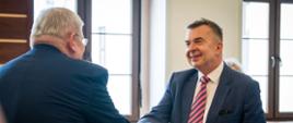 Minister Wieczorek stoi i podaje rękę mężczyźnie w ciemnoszarym garniturze, za nimi okno.