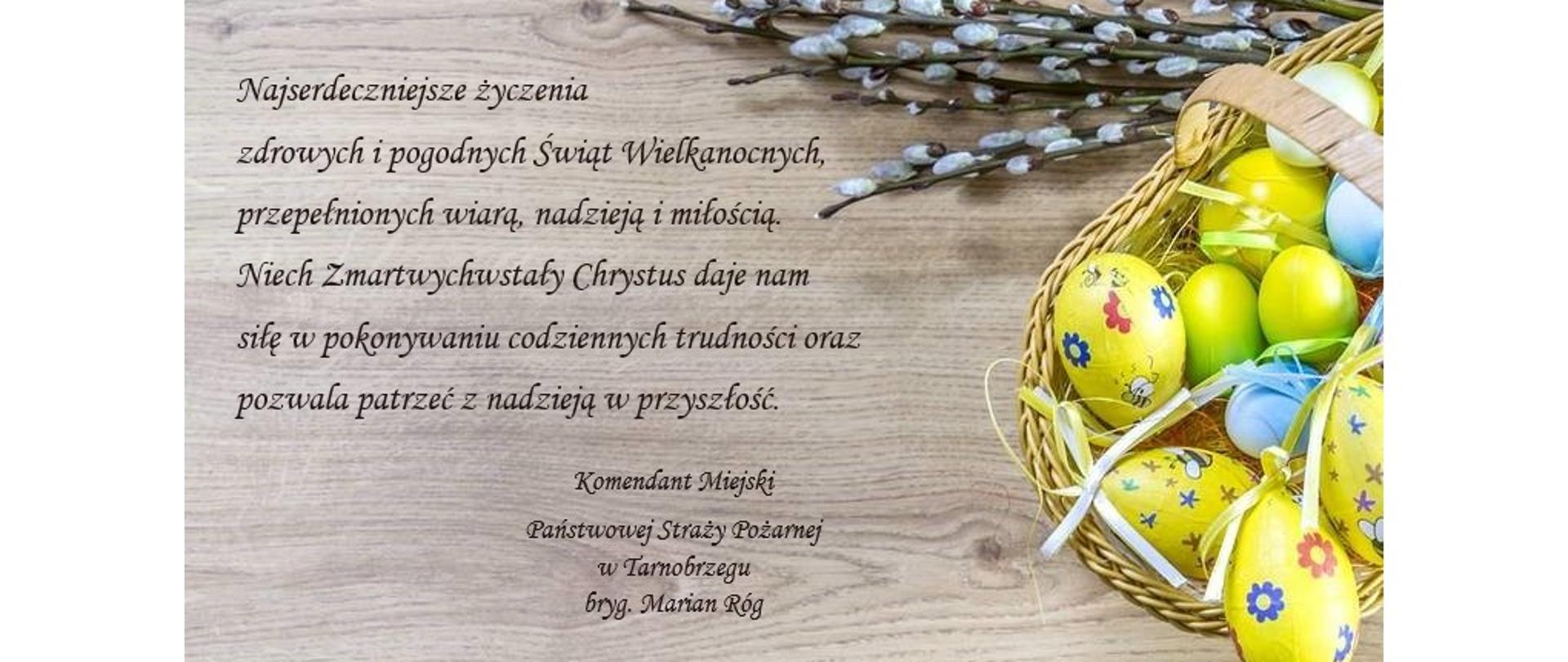 Na tle faktury deski koloru szarego, u góry widoczne pędy leszczyny (bazie), po stronie prawej widoczny koszyczek z pisankami. Po stronie prawej tekst życzeń wielkanocnych. 