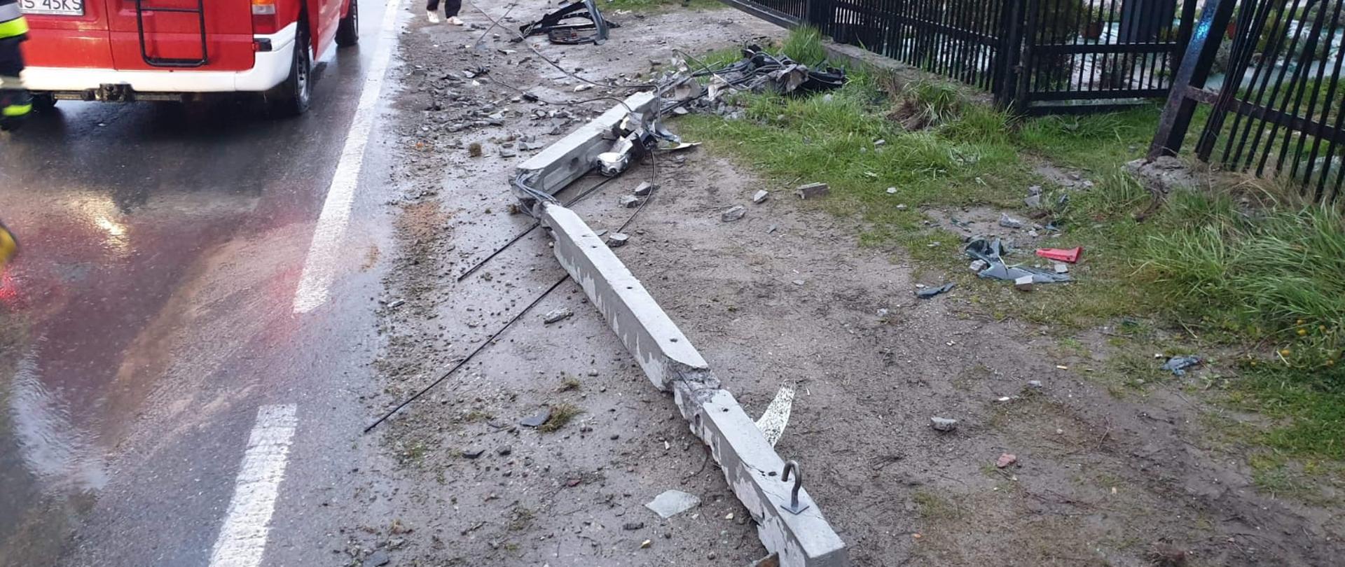 Połamany betonowy słup telekomunikacyjny na poboczu.Widać fragment samochodu pożarniczego oraz ogrodzenia posesji.