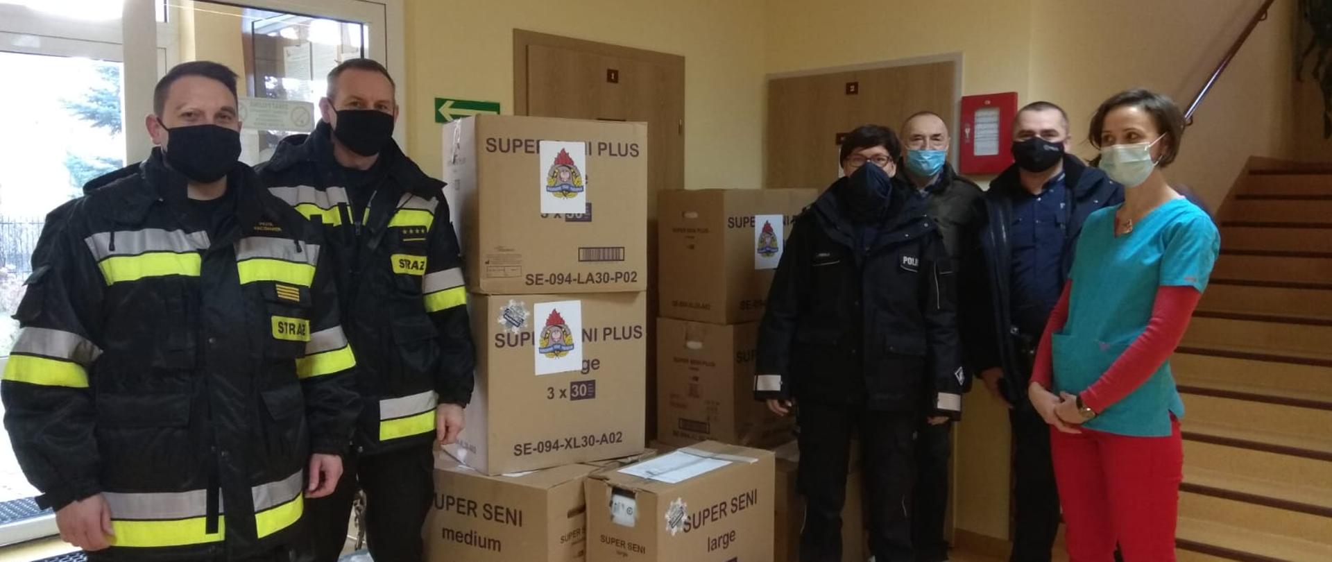 Na zdjęciu widać funkcjonariuszy straży pożarnej oraz policji z pracownikami hospicjum podczas przekazania paczek dla placówki