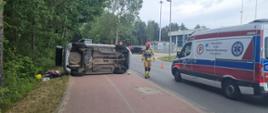 Na zdjęciu widoczny od lewej samochód leżący na boku, strażak w ubrany w ubranie koloru piaskowego z czerwonym hełmem oraz karetka pogotowia.