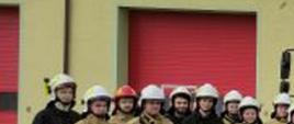 Zdjęcie przedstawia grupę 31 strażaków, którzy ukończyli kurs podstawowy dla OSP oraz w środku trzech funkcjonariuszy - oficerów PSP. Strażacy ochotnicy ubrani są w mundury bojowe, w hełmach białych, natomiast mundury w kolorze czarnym i część w kolorze piaskowy. Strażacy ustawieni w dwuszeregu, w półkolu na tle garaży Komendy Powiatowej PSP oraz na tle czerwonego samochodu ratowniczo – gaśniczego.