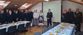 
Zdjęcie przedstawia spotkanie wielkanocne strażaków, które odbywa się w przestronnym pomieszczeniu z drewnianym sufitem. W centralnej części kadru, na wprost widza, widać dwa długie stoły pokryte białymi obrusami. Stoły są zastawione potrawami w miskach i na talerzach, a także dzbankami z sokiem pomarańczowym i szklankami.
Po lewej stronie stołów stoi grupa około dziesięciu strażaków w ciemnych mundurach galowych, z których część jest zwrócona w stronę stołów, a część patrzy w kierunku widza. Pomiędzy dwoma głównymi stołami, w tle, stoi grupa osób, w tym dwóch mężczyzn i dwie kobiety w ubraniach cywilnych, oraz jeden strażak w galowym mundurze, który stoi nieco z boku. Po prawej stronie zdjęcia, na dalszym planie, widać kolejną grupę strażaków w galowych mundurach. Na końcu pomieszczenia, na ścianie, zawieszony jest biały ekran projekcyjny.