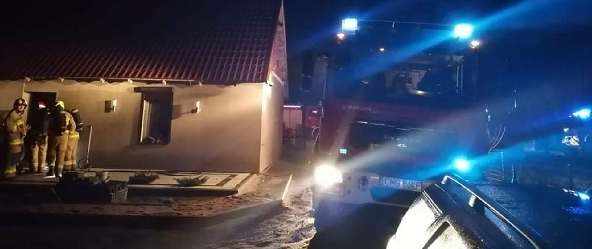 Po prawej stronie wóz strażacki, po lewej stronie budynek mieszkalny a przed nim 4 strażaków