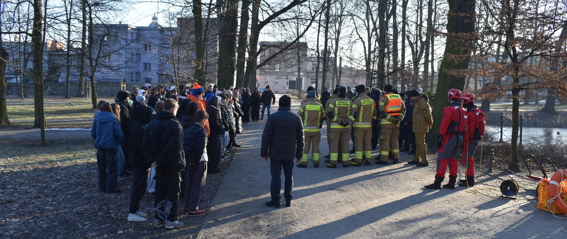 Nad stawem w parku miejskim trwa spotkanie edukacyjne dla uczniów dotyczące bezpiecznych ferii oraz bezpieczeństwa nad wodą w okresie zimowym. Strażacy oraz policjanci edukują uczniów szkół z powiatu krotoszyńskiego. Uczniowie zgromadzeni w grupie wysłuchują prelekcji 