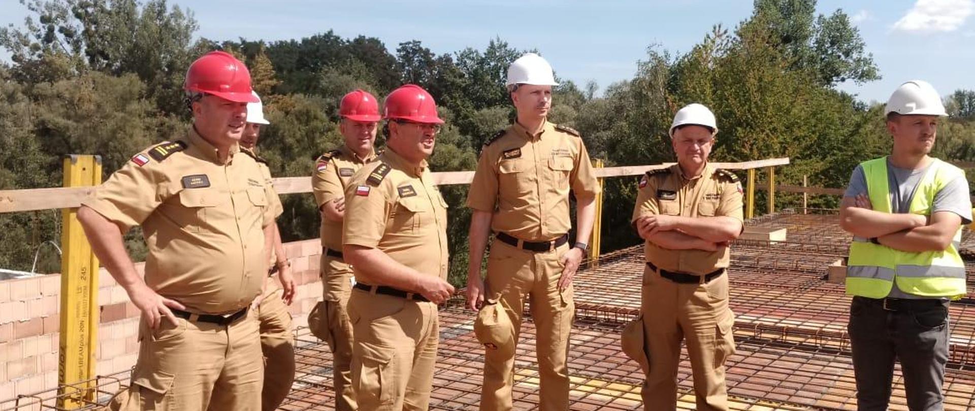 Grupa strażaków i pracowników budowy w kaskach ochronnych. Pod nogami widoczne zbrojenie. W tle drewniana barierka.