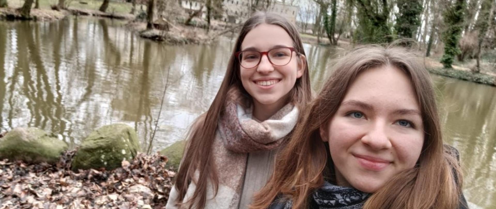 Zdjęcie kolorowe..Dwie dziewczyny pozują do zdjęcia. W tle staw i drzewa. W oddali jasny budynek z płaskim dachem.