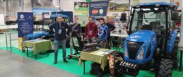 Pięciu mężczyzn stoi przy stoisku edukacyjnym na hali targowej. Obok nich znajduje się niebieski ciągnik rolniczy oraz symulator jazdy.