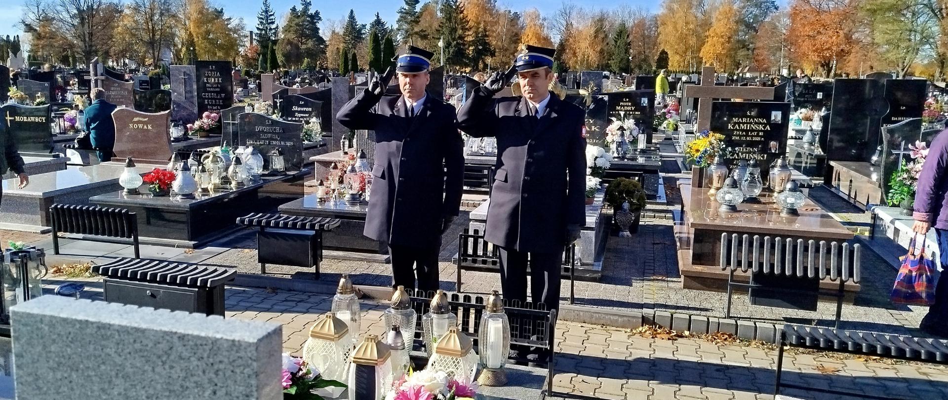 Na zdjęciu widać dwóch strażaków stojących przed grobem kolegi strażaka. Strażacy z stoją na baczność oddając hołd zmarłemu po uprzednim zapaleniu znicza. Jeden z nich ubrany jest w umundurowanie wyjściowe, drugi zaś w służbowe. Zdjęcie wykonane w porze południowej, mglistej. W tle nagrobki.