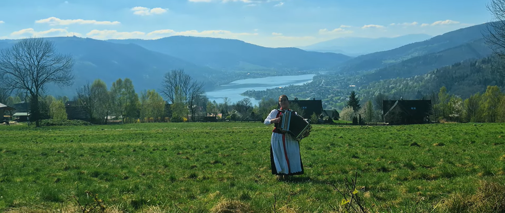 Uczennica w tradycyjnym stroju grająca na akordeonie na łące, w tle domy, jezioro i górzysty krajobraz.