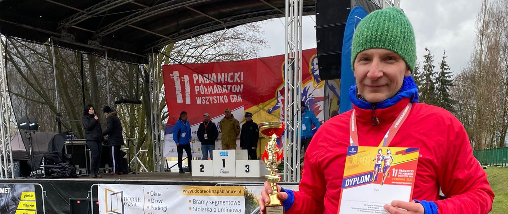 Na zdjęciu mł. ogn. Miłosz Michalak, który właśnie otrzymał pamiątkowy dyplom i puchar za zajęcie 3 miejsca w kategorii PSP w XI Pabianickim Półmaratonie. W tle scena z podium.