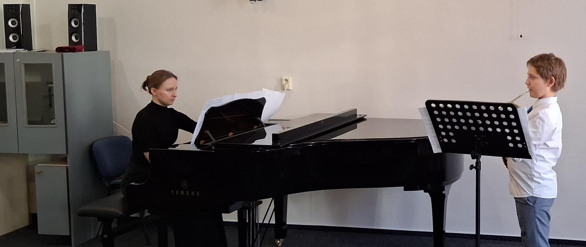 Z prawej strony waltornista w galowym stroju, z lewej przy fortepianie pianistka. 