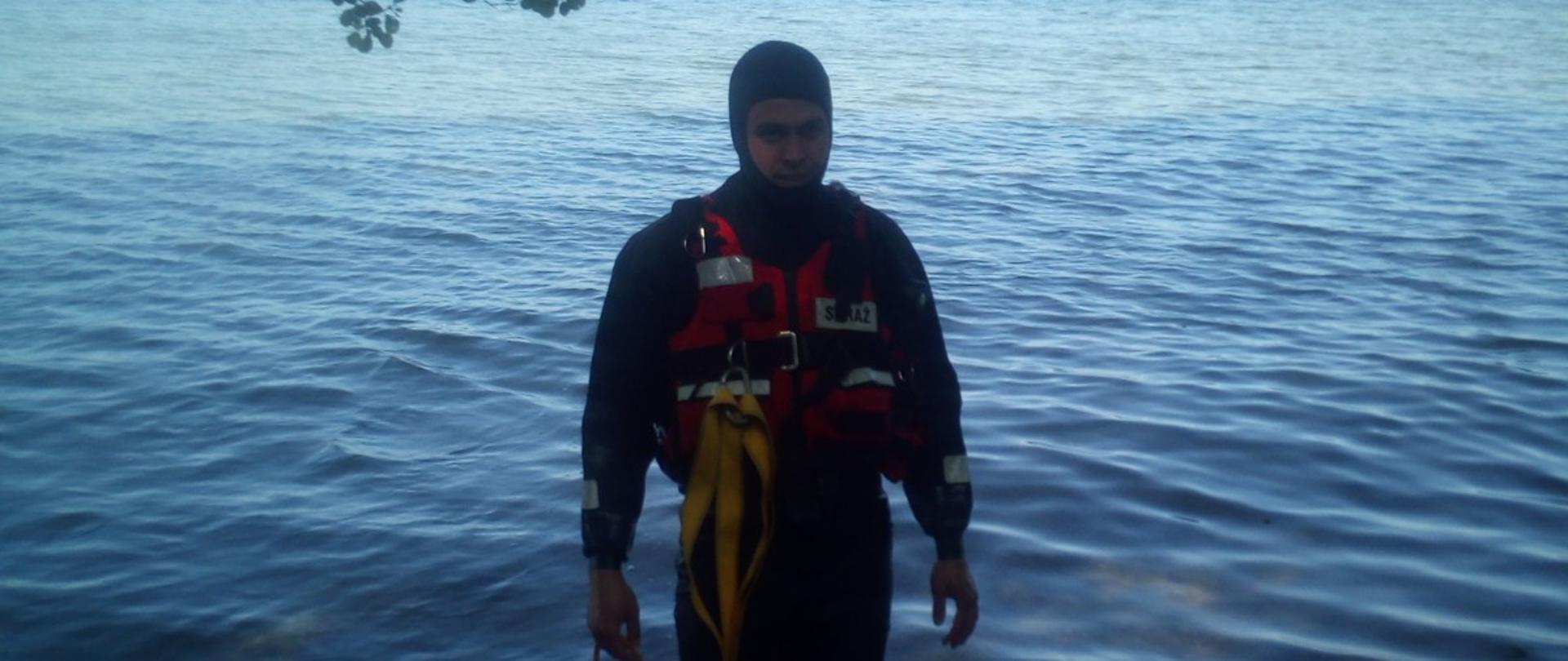 Strażak ubrany w specjalne ubranie stoi w wodzie do łydek. Zabezpieczony jest linką ratowniczą. W oddali widać 2 brzeg jeziora. 