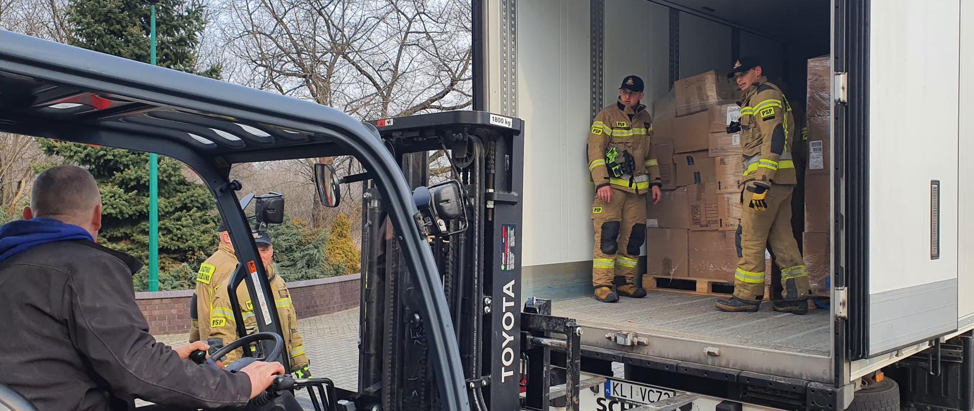 Na pierwszym planie wózek widłowy w którym siedzi mężczyzna . Wózek podjeżdża do dużej naczepy na której stoją dwaj strażacy i rozładowują pudła. Naczepa i wózek stoją na placu wyłożonym kostką. W tle widać drzewa.