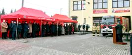 Na pierwszym planie dwa czerwone namioty pod którymi stoją zaproszeni goście biorący udział w przekazaniu samochodu pożarniczego. Samochód stoi na tle budynku remizy nad nim napis Ochotnicza Straż Pożarna Zagórów 1899 z boku stoi dwóch strażaków ubranych w ubrania specjalne koloru piaskowego przy mównicy strażak w mundurze. 
