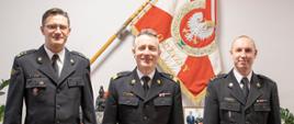 Zdjęcie pamiątkowe. Trzech mężczyzn w mundurach na tle sztandaru Komendy Głównej Państwowej Straży Pożarnej
