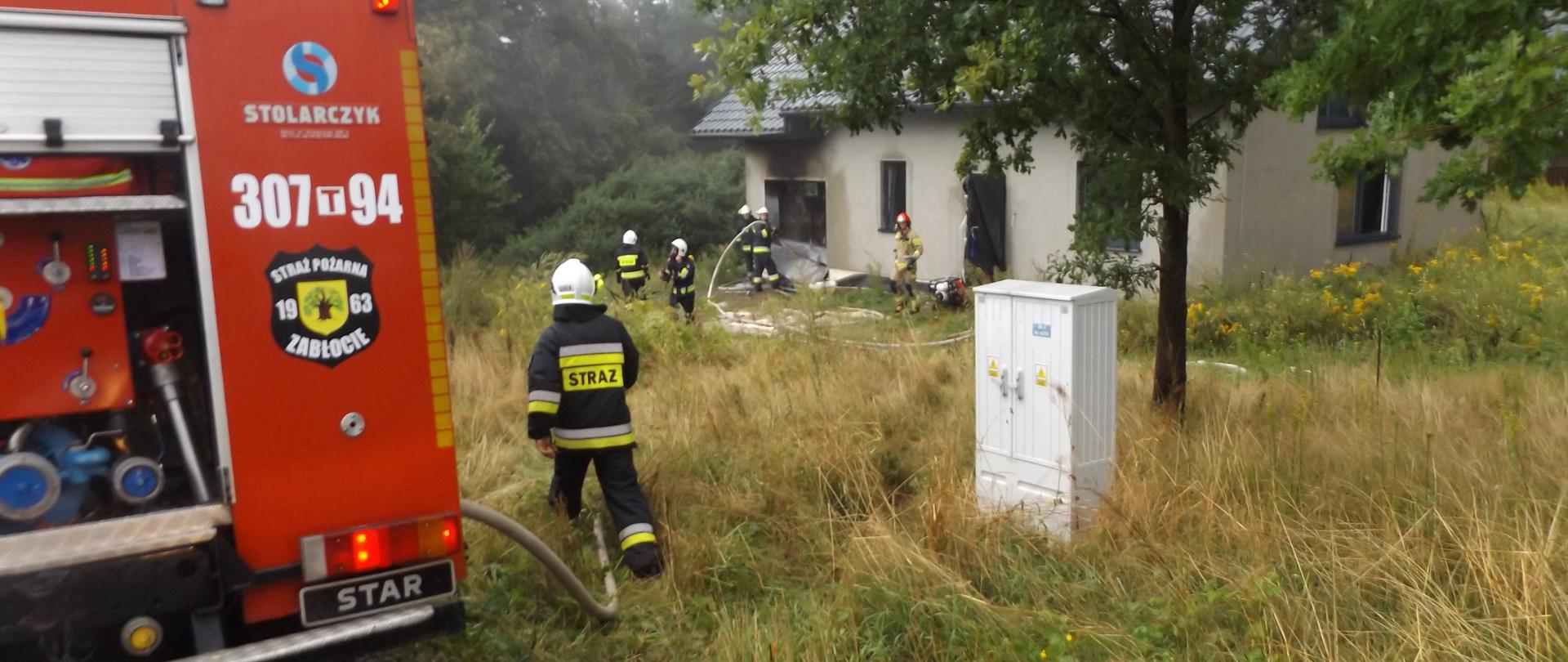 Zdjęcie przedstawia samochód strażacki przed domem w którym doszło do pożaru. Za samochodem widać strażaków gaszących pożar.