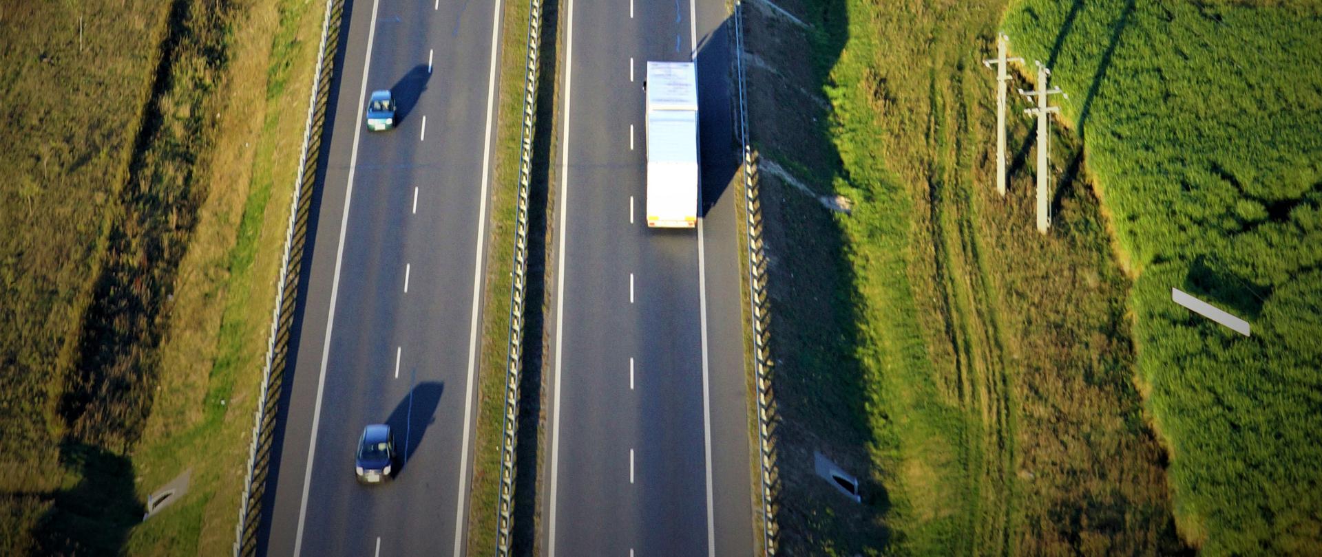 Droga krajowa - zdjęcie ilustracyjne. Fotografia przedstawia dwujezdniową drogę krajową z ruchem pojazdów poprowadzoną na nasypie.