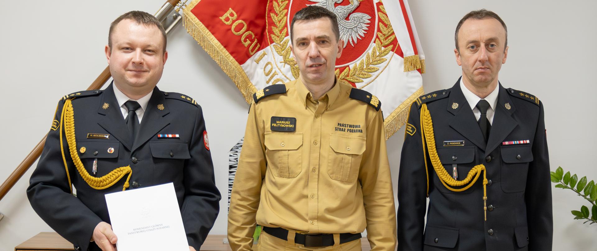 Komendant główny PSP pozuje do zdjęcia z nowo powołanym zastępcą lubelskiego komendanta wojewódzkiego PSP oraz z lubelskim komendantem wojewódzkim PSP. W tle na ścianie znajduje się sztandar. 