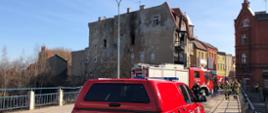 Zdjęcie przedstawia miejsce pożaru, na pierwszym planie samochody pożarnicze, w tle widać budynek, opalone i wybite okna, widoczne działanie ognia.
