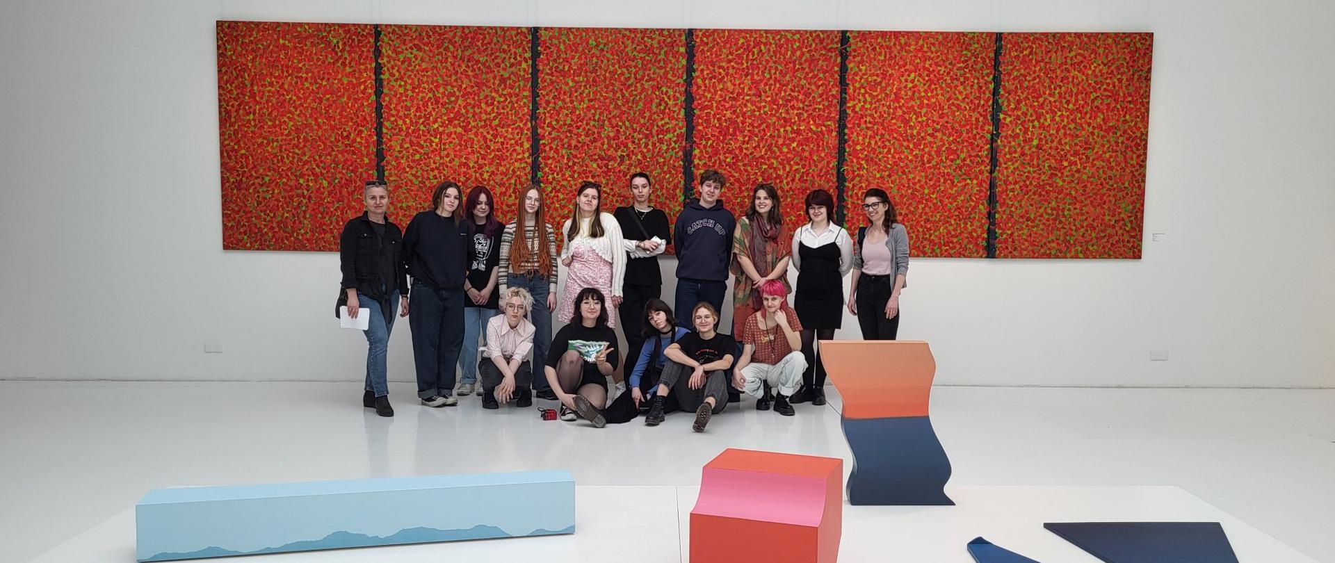 Uczestnicy wycieczki w muzeum, za nimi obraz z czerwonymi plamami, przed nimi geometryczne obiekty leżące na podłodze