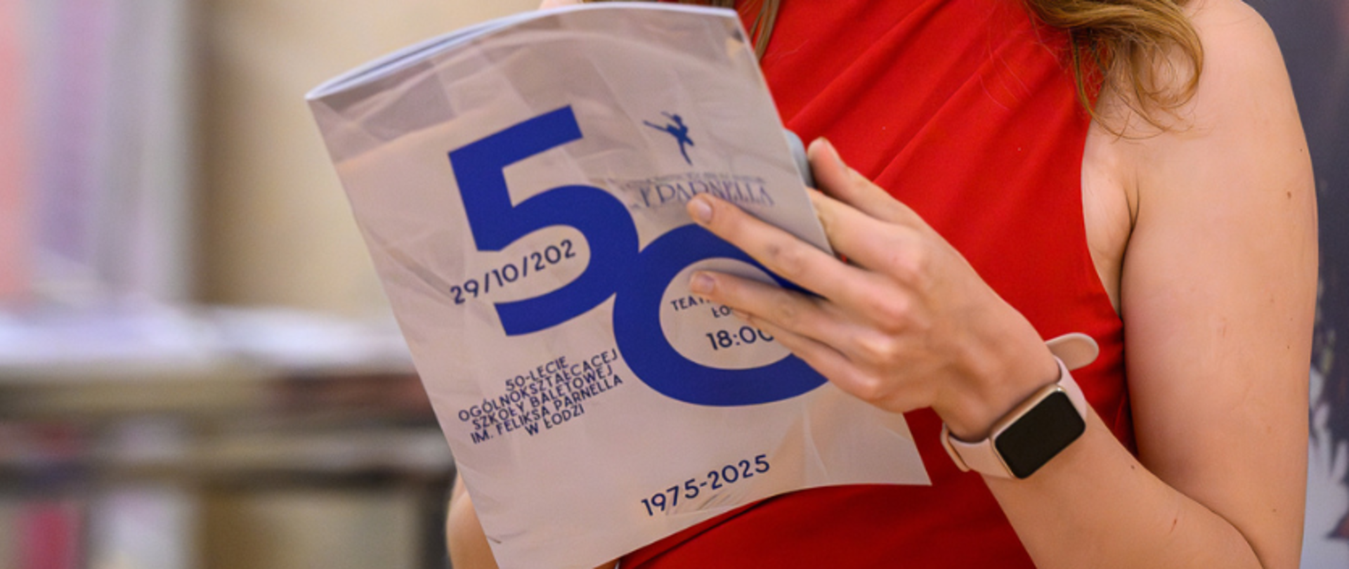 zdjęcie: na zdjęciu postać kobiety w czerwonej sukience, która trzyma w ręku folder jubileuszowy 50-lecia Ogólnokształcącej Szkoły Baletowej im. Feliksa Parnella