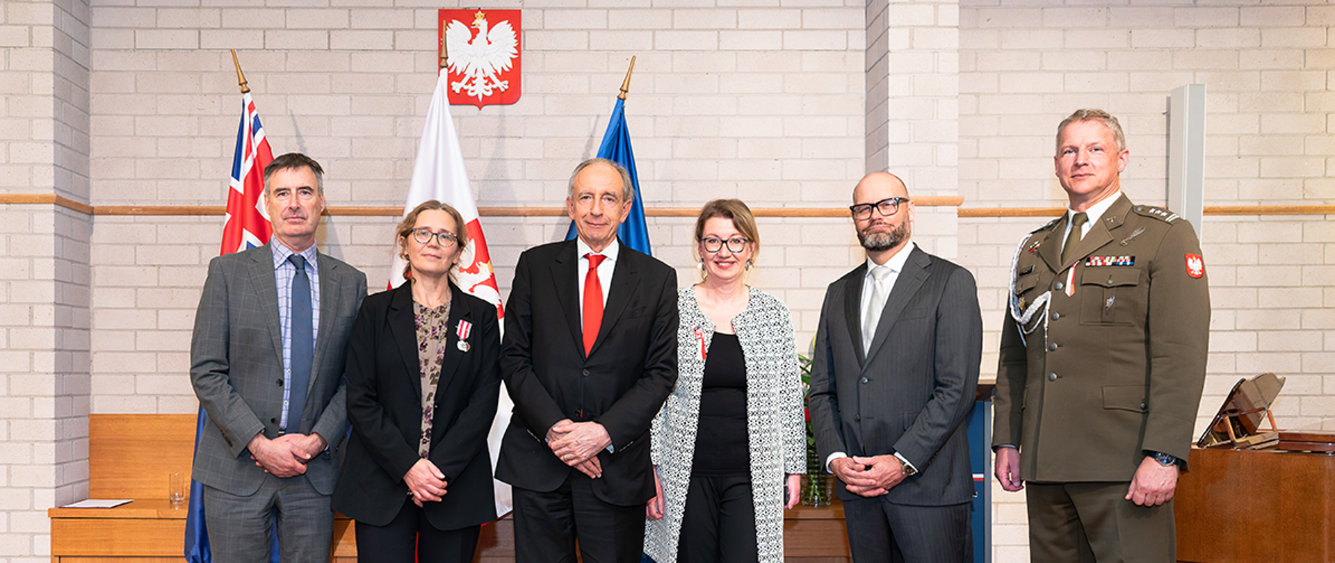 Grupa osób na zdjęciu stoi na tle flag Australii, Polski i Unii Europejskiej