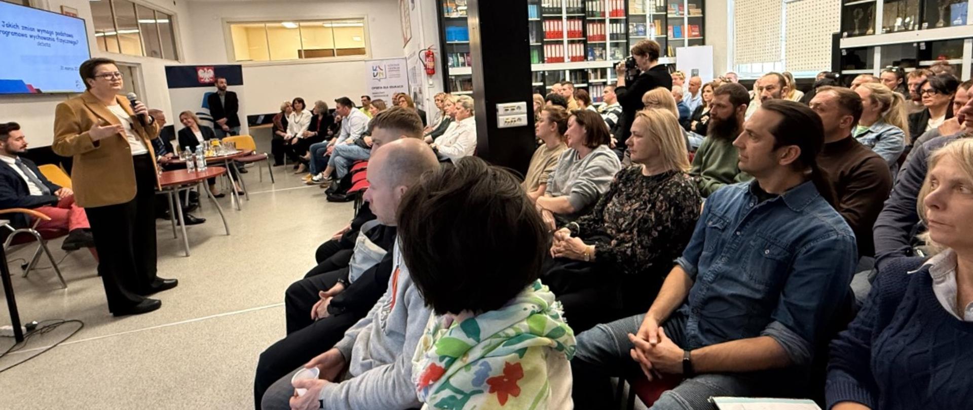 Wiceminister Katarzyna Lubnauer przemawia do mikrofonu na spotkaniu podczas spotkania z nauczycielami. Nauczyciele siedzą i słuchają wiceminister. 