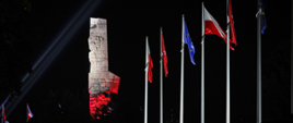 Pomnik podświetlony w kolorze flagi Polski obok na masztach wiszą flagi Polski Miasta Gdańska oraz Unii Europejskiej.