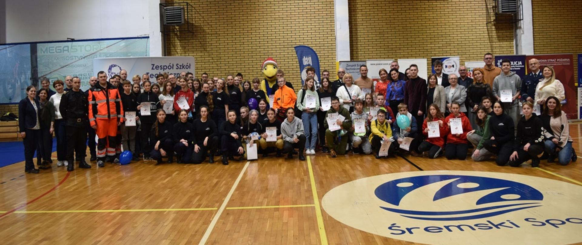 Zdjęcie grupowe uczestnicy, goście organizatorzy sędziów uczniowie z przodu klęczą wszyscy na płycie hali śremskiego sportu